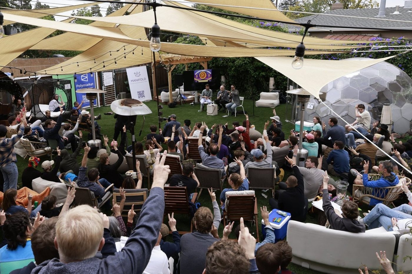 A large crowd at an outdoor conference session, many with hands raised.