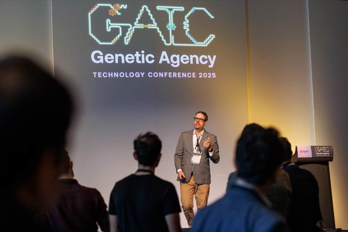 Misha speaking on stage at the Genetic Agency Technology Conference, seen past the silhouettes of standing attendees.