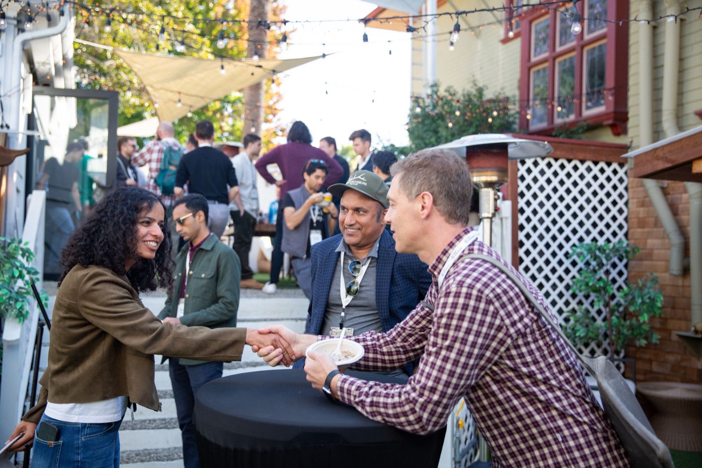 Two attendees shaking hands across a high table at an outdoor conference reception, with a third looking on.