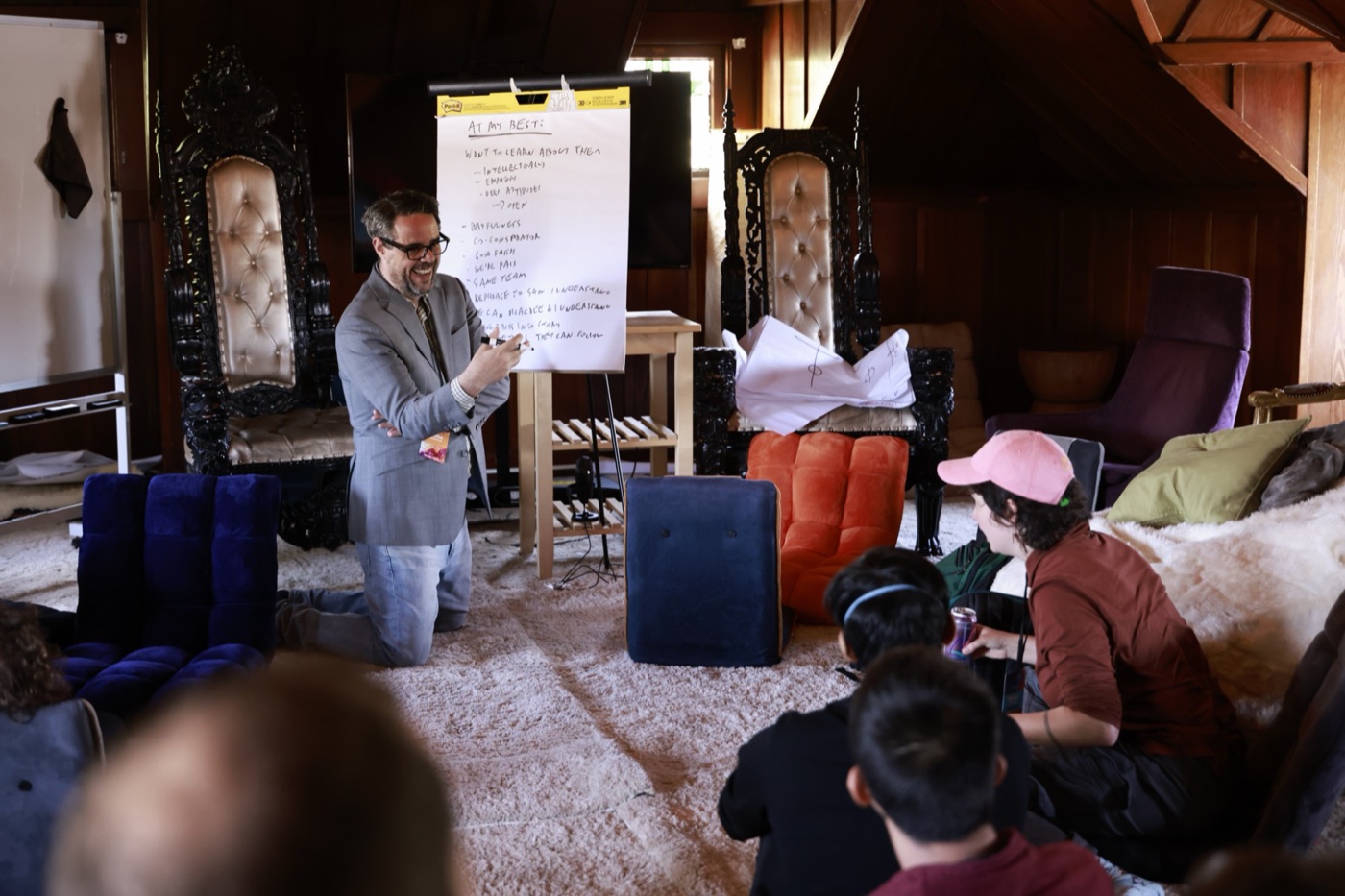 Misha kneeling beside a flip chart, laughing as he leads a workshop for people seated on floor cushions.