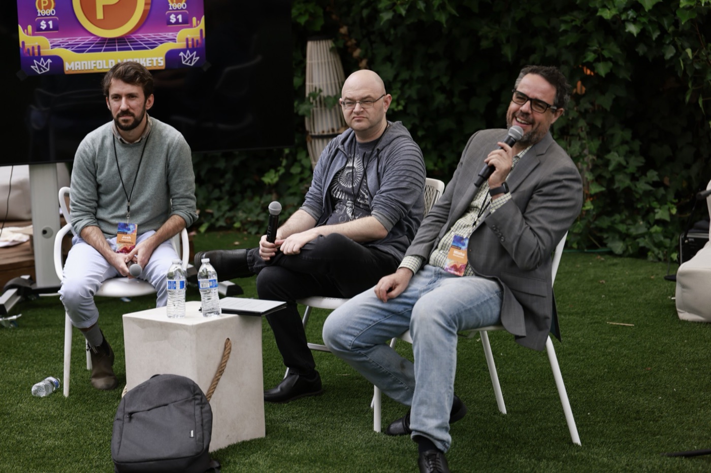 Misha laughing into a microphone while seated on an outdoor panel with two other speakers.
